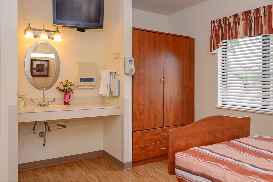 A nursing care center bedroom featuring a wooden bed with a striped bedspread, a wooden wardrobe, a window with blinds and a red and white valance, a wall-mounted TV, a sink with a mirror above it, a paper towel dispenser, and a hand sanitizer dispenser. There is also a small vase with flowers on the sink counter.
