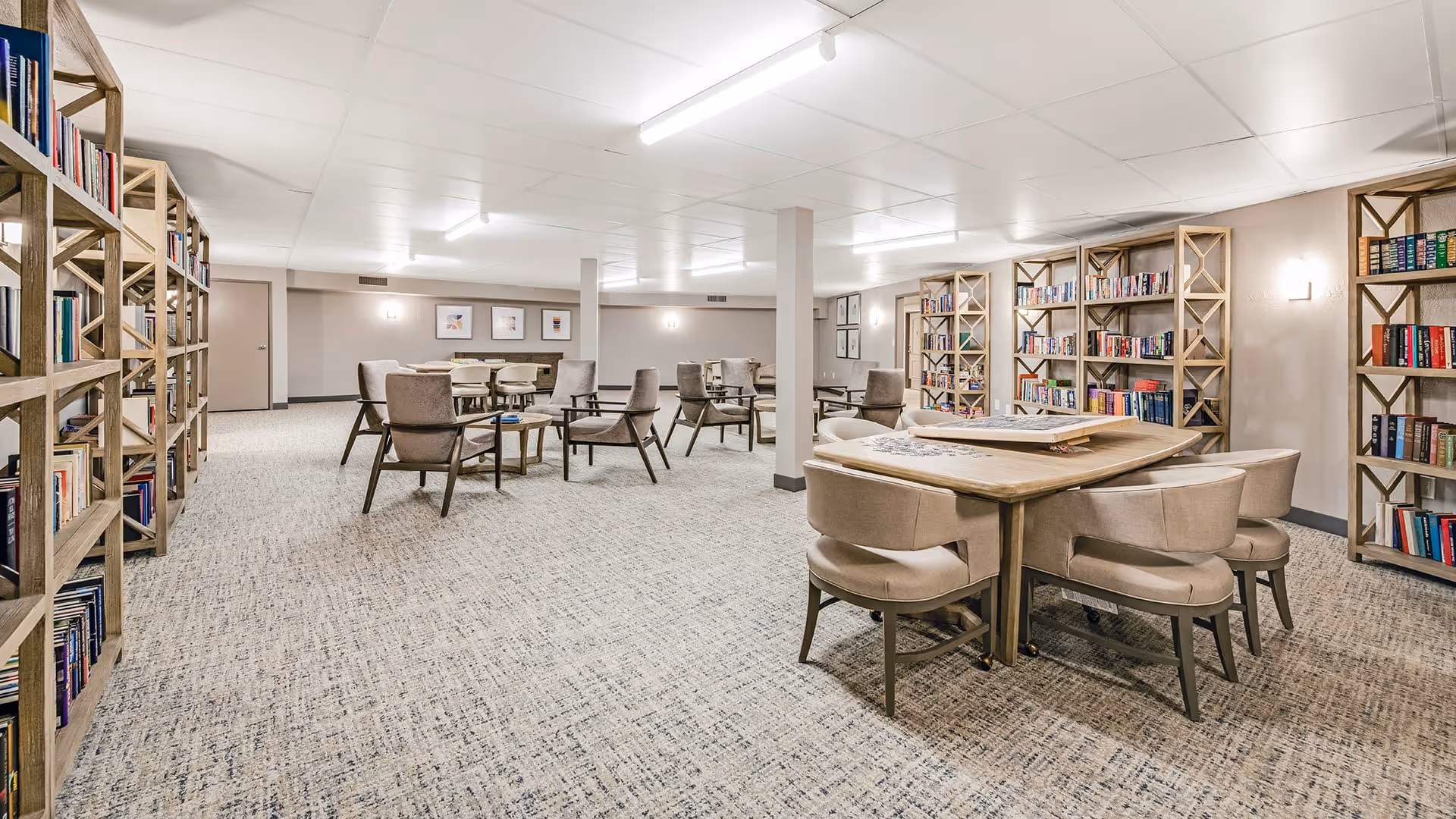 A spacious, well-lit room with multiple bookshelves filled with books along the walls. Several tables and comfortable chairs are arranged throughout the room, suggesting a communal reading or activity area.