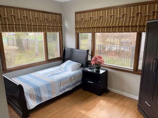 A small bedroom with a single bed covered in a blue and white striped quilt, a black nightstand with a pink flower pot and a lamp, large windows with brown patterned blinds, and a dark wooden wardrobe. Outside the windows, a fenced garden area is visible.