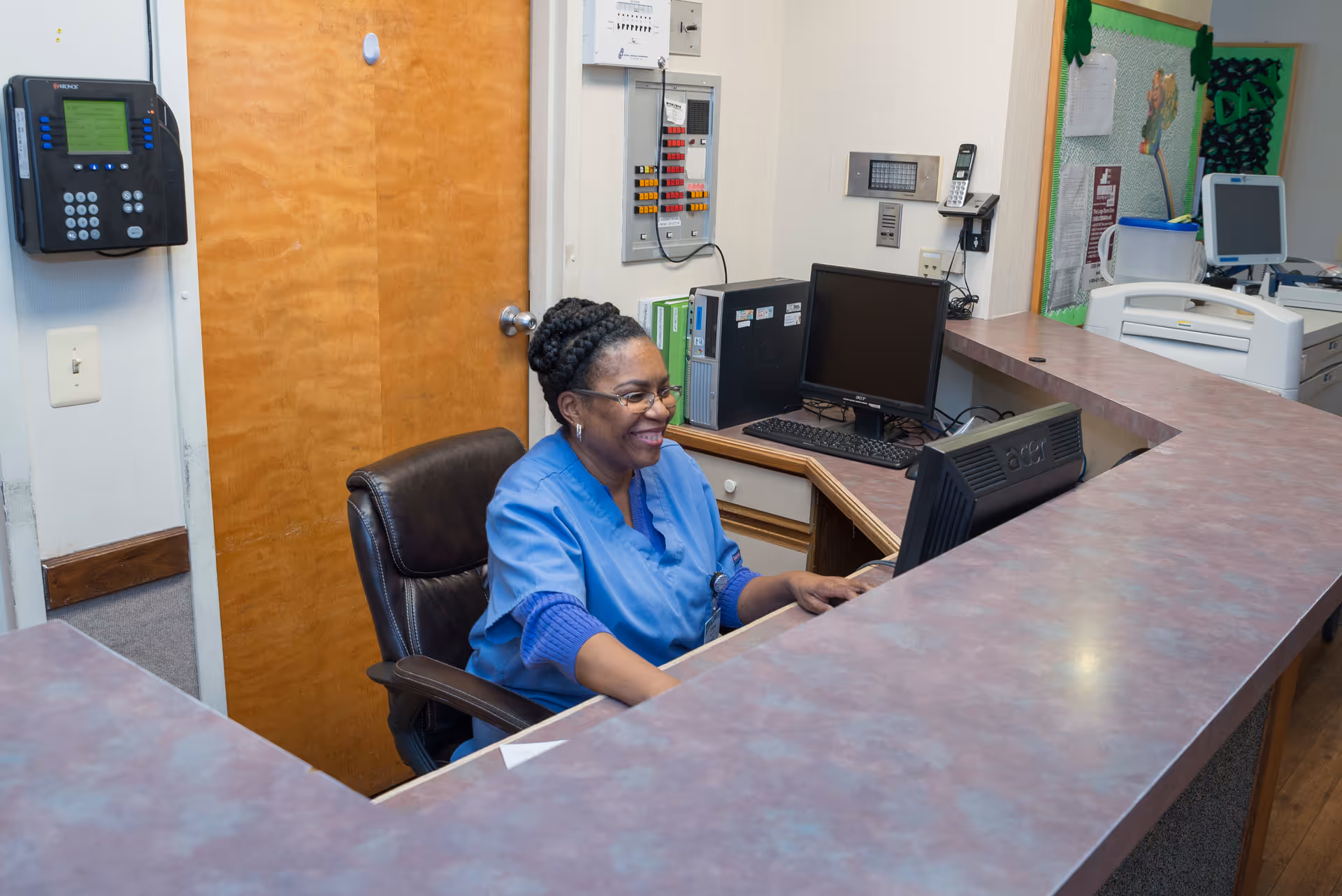 A smiling woman in blue scrubs sitting at a reception desk with computer monitors and office equipment in the background.