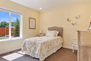 A bright bedroom with a single bed covered in a floral bedspread, a nightstand with a lamp, a framed picture on the wall, three decorative butterflies mounted on the wall, a wooden dresser, and a window showing a fenced backyard with trees.