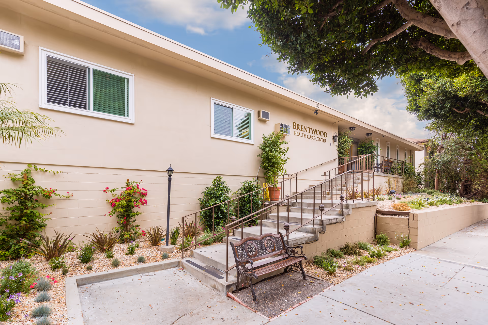 Exterior view of Brentwood Health Care Center showing a beige building with windows, a staircase with metal railings leading to the entrance, a bench at the base of the stairs, and landscaped plants and trees around the walkway under a partly cloudy sky.
