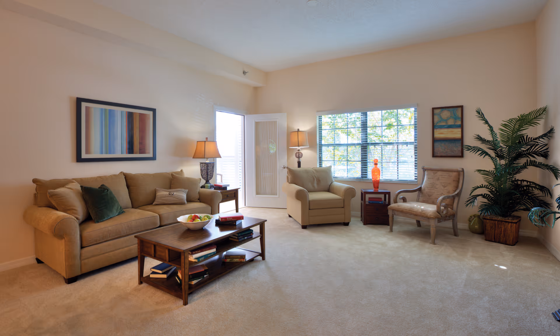 A cozy living room with beige walls and carpet, featuring a beige sofa with multiple cushions, a wooden coffee table with books and a bowl of fruit, a beige armchair, a wooden side table with a lamp, a decorative chair, a large window with blinds, two framed artworks on the walls, and a potted plant in the corner.
