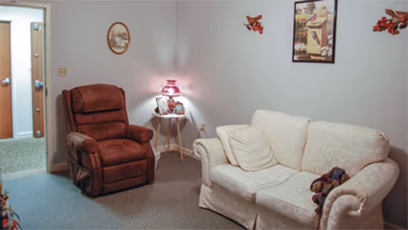 A cozy living room with a brown recliner chair and a white patterned sofa. A small round side table with a pink lamp and framed photos is placed between the chairs. The walls are decorated with a round picture and a framed artwork featuring birds and flowers.