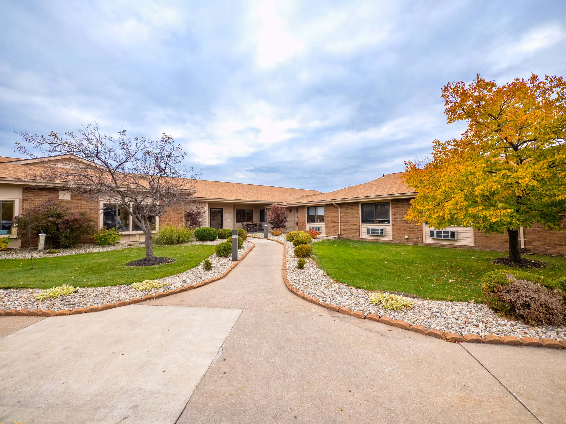 Exterior view of Edgewood Manor Rehabilitation and Healthcare Center showing a single-story brick building with a tan roof. There is a paved walkway leading to the entrance, bordered by green grass, small bushes, and trees with autumn foliage under a cloudy sky.