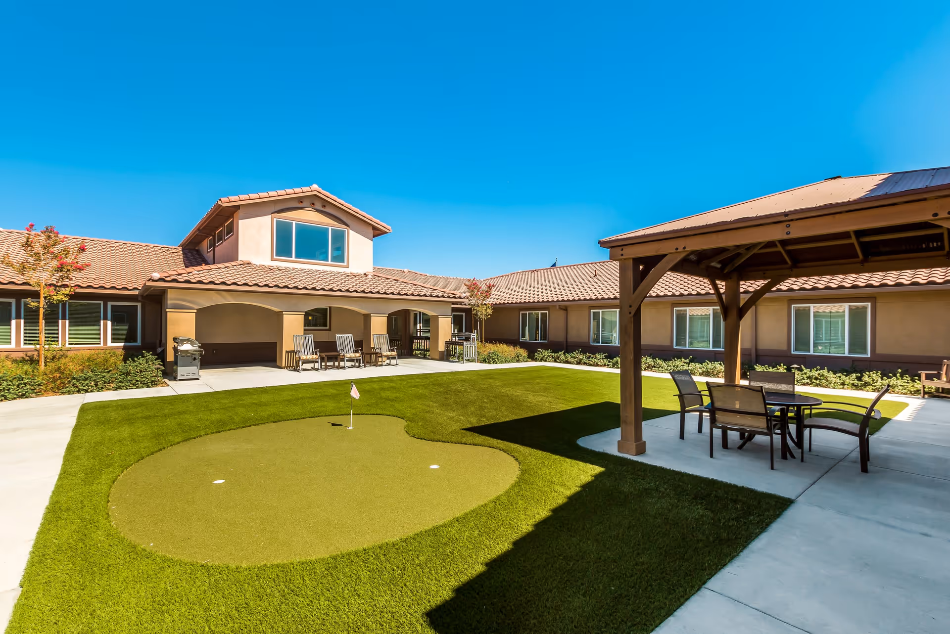 Outdoor courtyard area at Vineyard Place featuring a small putting green with a flag, surrounded by well-maintained grass and concrete walkways. There is a covered seating area with a table and chairs on the right side, and a building with a tiled roof and multiple windows in the background under a clear blue sky.