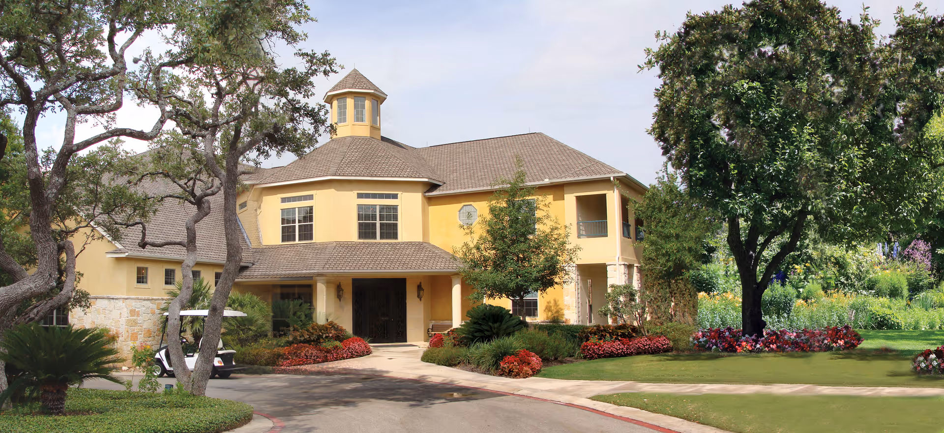 Exterior view of a large, yellow retirement community building with a turret, surrounded by well-maintained landscaping including trees, bushes, and flower beds. A golf cart is parked near the entrance.