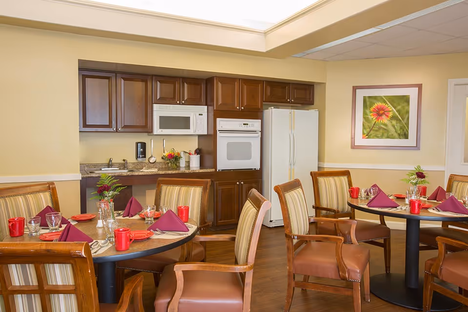 Dining area with round tables set with red mugs and purple napkins in front of a kitchenette with wooden cabinets, microwave, oven, and refrigerator.