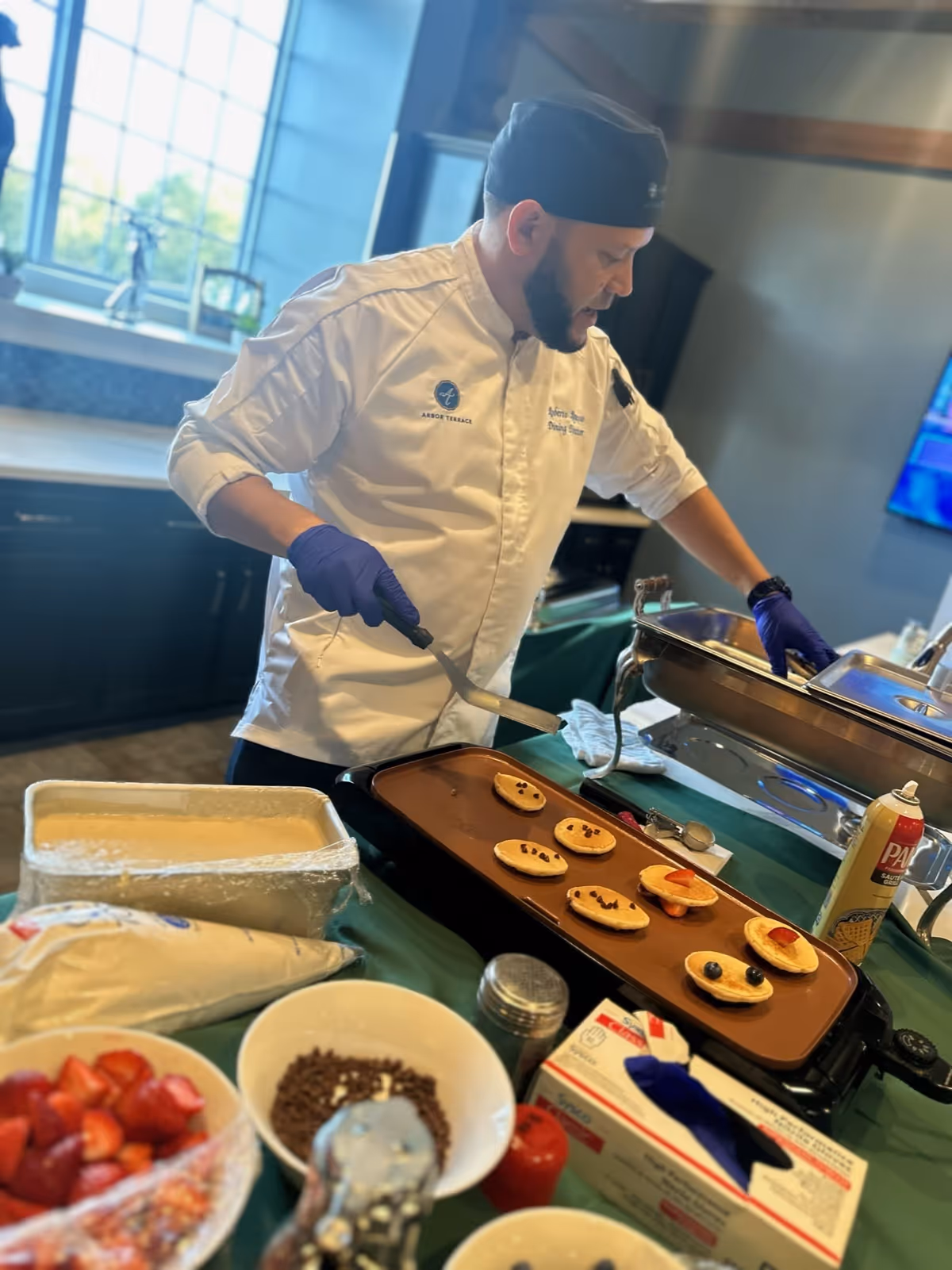A chef wearing a white Arbor Terrace Mountainside uniform and black hat is cooking small pancakes on a griddle. The pancakes are decorated with fruit and chocolate chips to create smiley faces. Various bowls of ingredients, including strawberries, chocolate chips, and whipped cream, are on the table in front of him. The setting appears to be a kitchen or dining area with a window in the background.