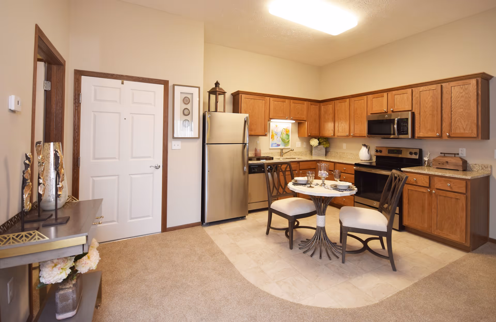 A cozy kitchen area in a retirement community featuring wooden cabinets, stainless steel refrigerator, dishwasher, microwave, and stove. A small round dining table with two chairs is set with plates and glasses. The floor transitions from carpet to tile in the kitchen area, and there is a decorative console table with vases and flowers near a closed white door.
