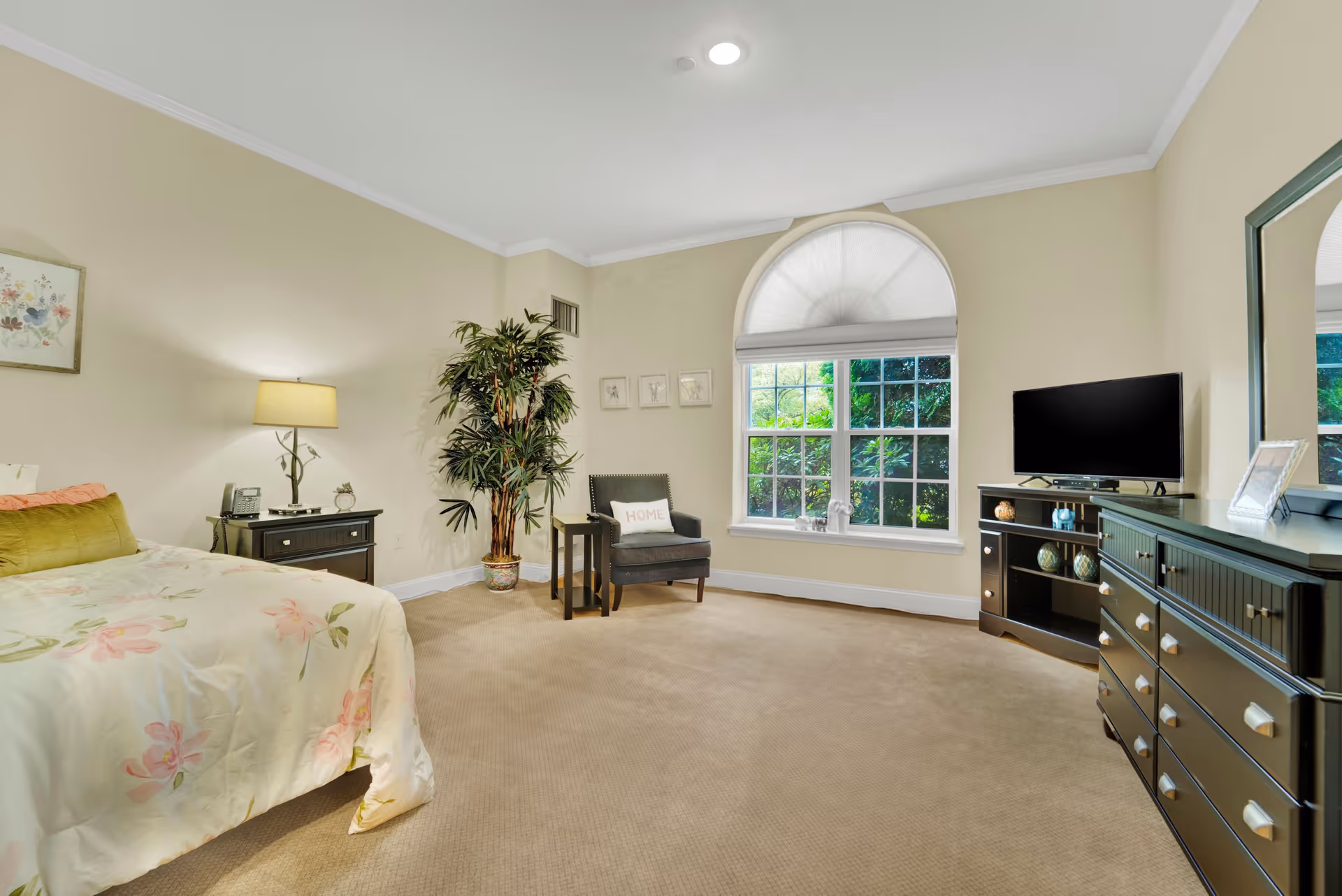 A bright bedroom with beige walls and carpet, featuring a bed with floral bedding on the left, a black nightstand with a lamp and phone, a potted plant, a gray armchair with a 'HOME' pillow, a large arched window with a white shade, and a black dresser with a mirror and a TV on top.