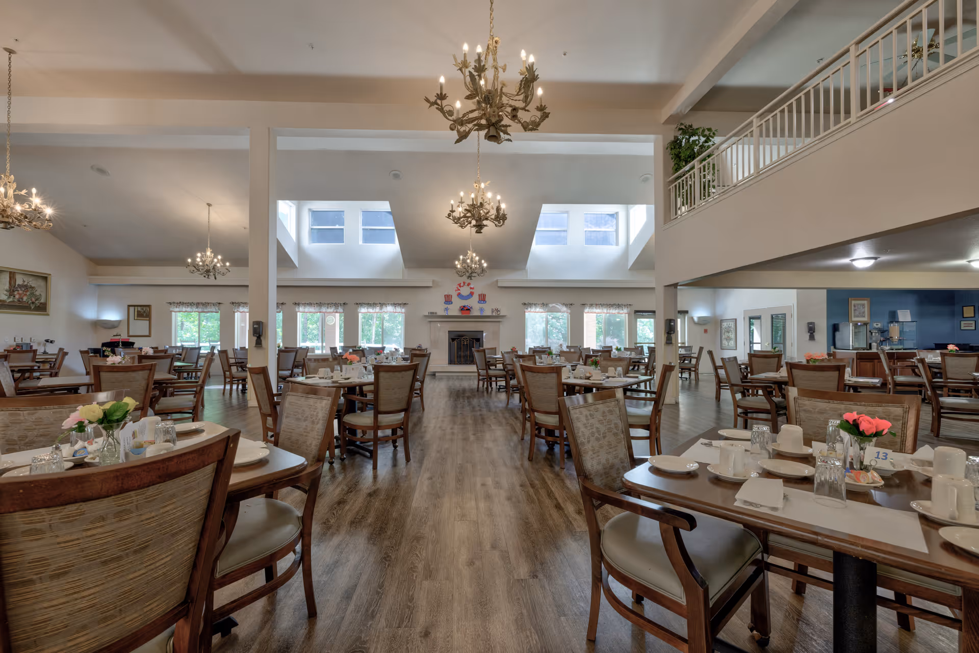 Spacious dining room with multiple wooden tables and chairs arranged neatly. Tables are set with plates, cups, napkins, and small flower arrangements. The room features large windows letting in natural light, chandeliers hanging from the ceiling, and a fireplace at the far end. There is a second-floor balcony visible on the right side.