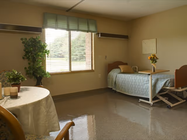 A simple, clean bedroom in a senior living facility with a single bed covered in a light blue bedspread, a small table with a vase of sunflowers, a round table with a white tablecloth and some candles, a potted plant near the window, and a large window with blinds letting in natural light.