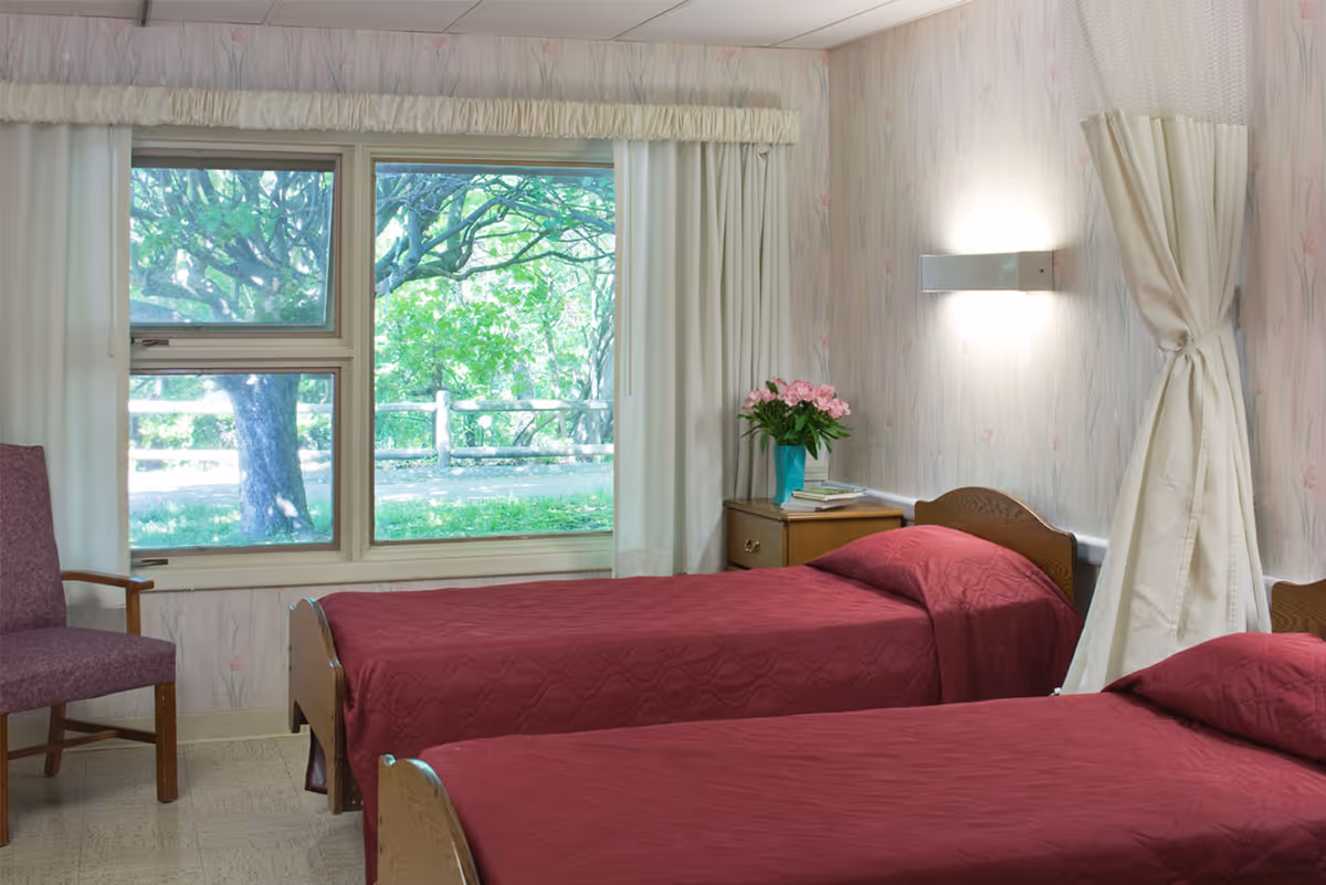 A room with two single beds covered in maroon bedspreads, a wooden nightstand with a vase of pink flowers and books, a purple armchair, and a large window showing green trees outside. The walls have light-colored wallpaper with a subtle pattern, and a white curtain is tied back on one side.