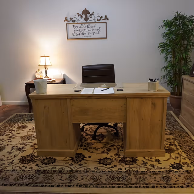 A wooden desk with a black office chair behind it, placed on a patterned rug. On the desk are papers, a pen, a small container with pens, and a white bucket. To the left of the desk is a side table with a lit lamp and decorative items. A framed inspirational quote hangs on the wall above the desk, and a tall green plant is positioned to the right side of the desk.