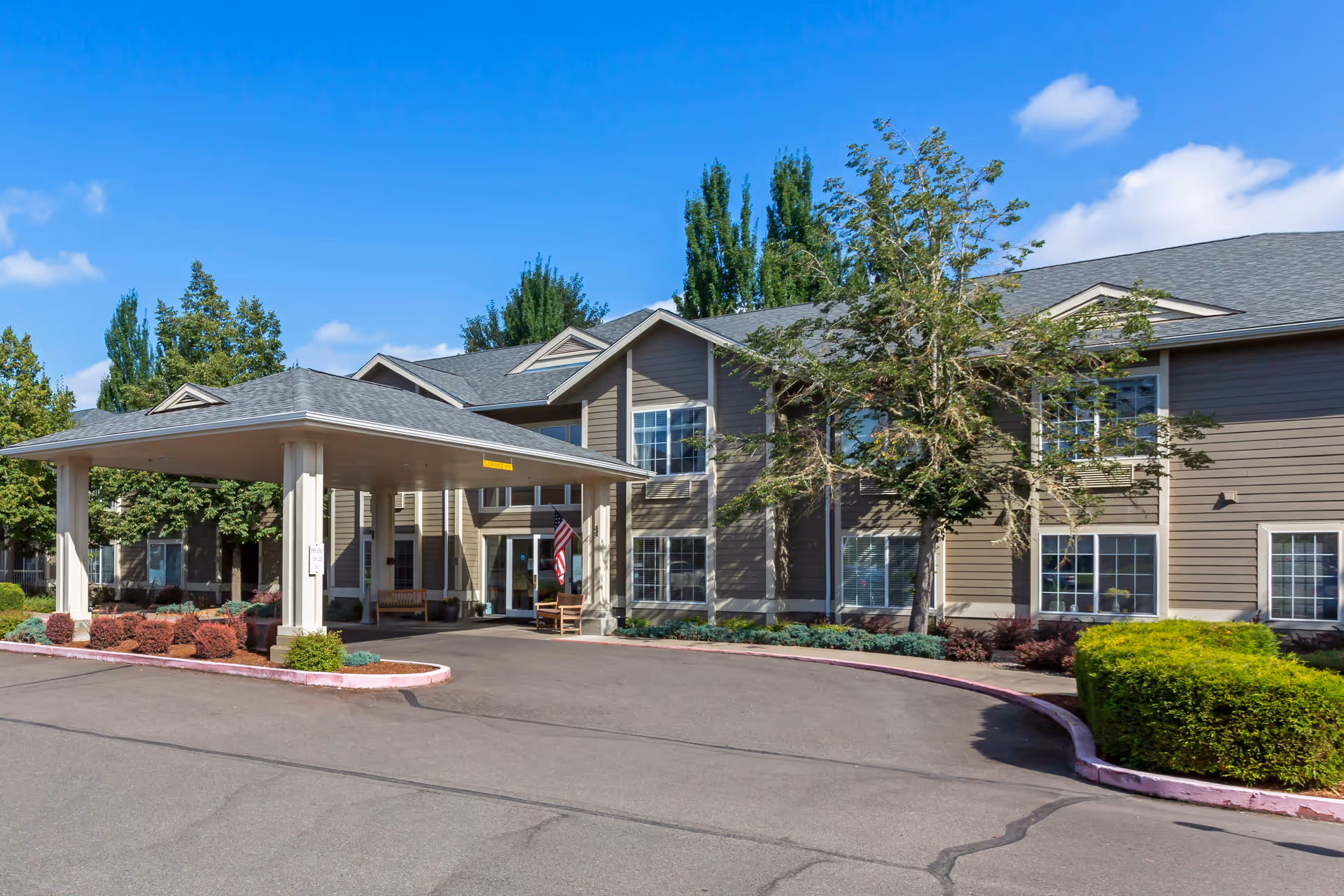 Exterior view of a two-story senior living facility building with a covered entrance, surrounded by trees and shrubs under a clear blue sky.