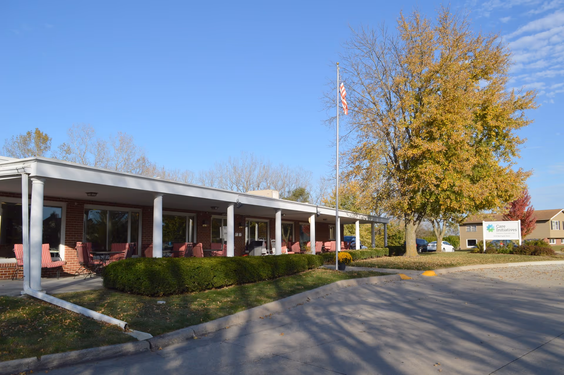 Exterior view of a single-story brick building with a covered porch featuring several red cushioned chairs and tables. There is a flagpole with an American flag in front of the building, surrounded by green bushes and a large tree with autumn-colored leaves. A sign near the tree reads 'Care Initiatives'. The sky is clear and blue.