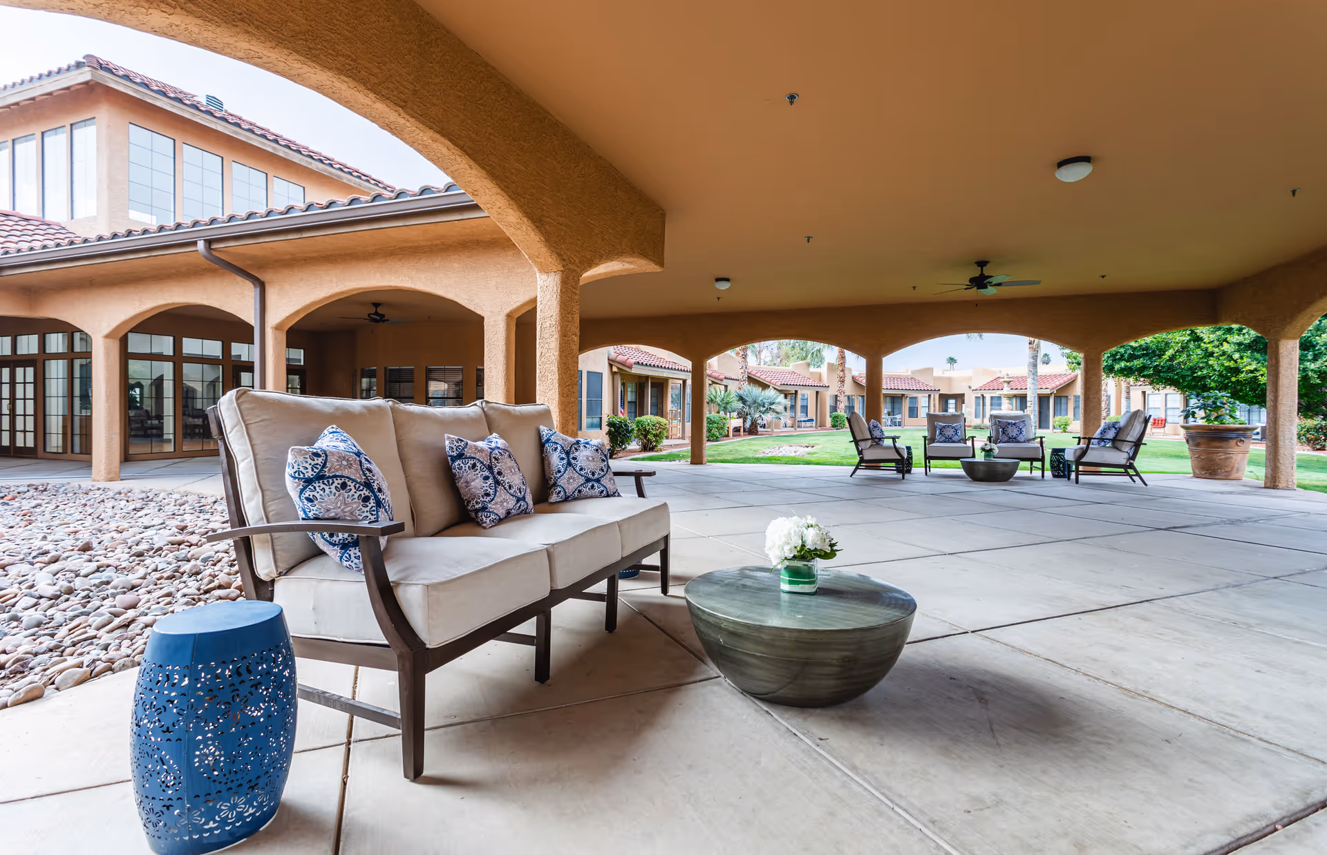 Covered outdoor patio area at a senior living facility with cushioned seating, decorative pillows, a round coffee table with a small flower arrangement, and additional seating in the background. The patio is surrounded by a building with arches and large windows, and there is a landscaped garden with grass and trees visible beyond the patio.