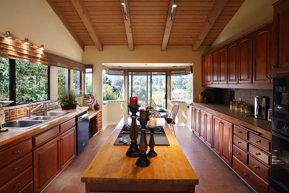 A spacious kitchen with wooden cabinets and a wooden island countertop featuring three decorative candle holders. The kitchen has a double sink under a large window with a view of greenery outside. In the background, there is a dining area with a round table and chairs near large windows with bamboo blinds, allowing natural light to fill the space. The ceiling has exposed wooden beams.