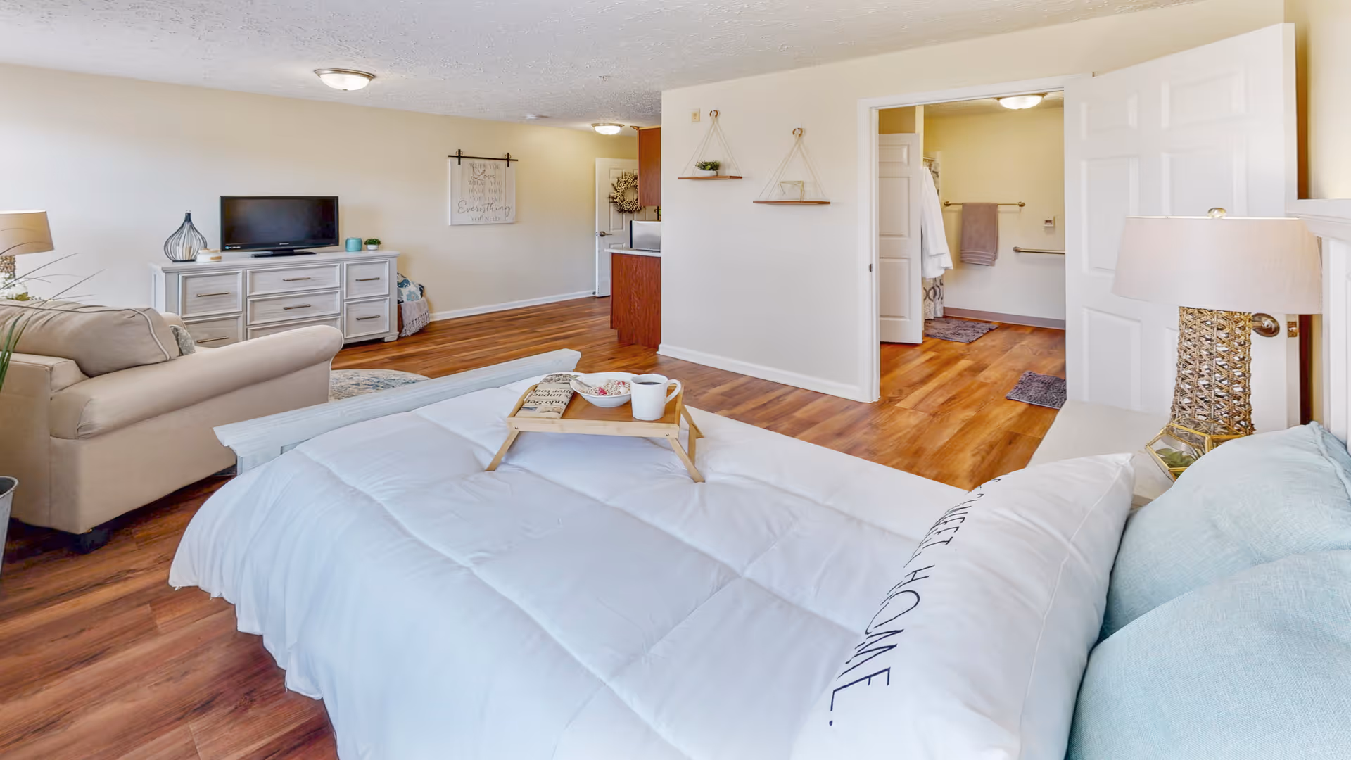 A bright and spacious bedroom with a large bed covered in a white comforter and light blue pillows. A wooden tray with a book, bowl, and cup sits on the bed. To the left, there is a beige armchair and a white dresser with a TV on top. The room has wooden flooring and white walls, with an open doorway leading to a bathroom area with towels and a closet visible.