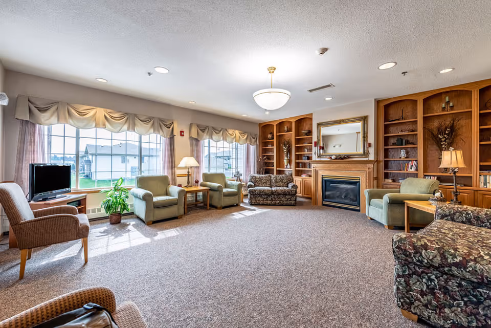 A spacious living room with large windows allowing natural light to fill the space. The room features multiple armchairs and sofas arranged around a central area, a wooden fireplace with a mirror above it, built-in wooden bookshelves, a television on a stand, and several lamps providing additional lighting.