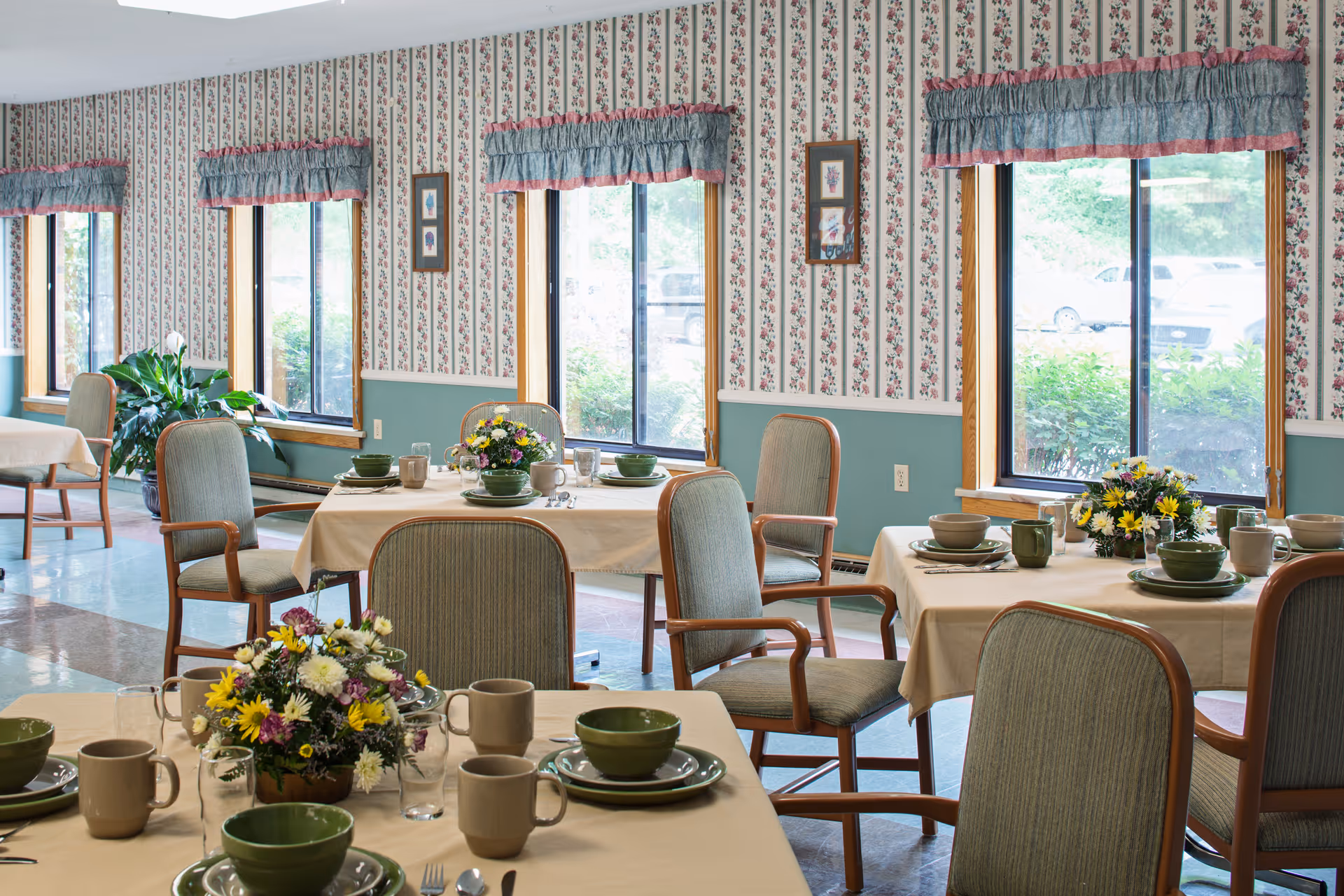 A dining room in a senior living facility with tables set for meals. Each table has beige tablecloths, green bowls, beige mugs, plates, and silverware. There are floral centerpieces on the tables. The room has large windows with floral-patterned curtains and wallpaper with a floral design. Several chairs with wooden arms and green upholstery are arranged around the tables.
