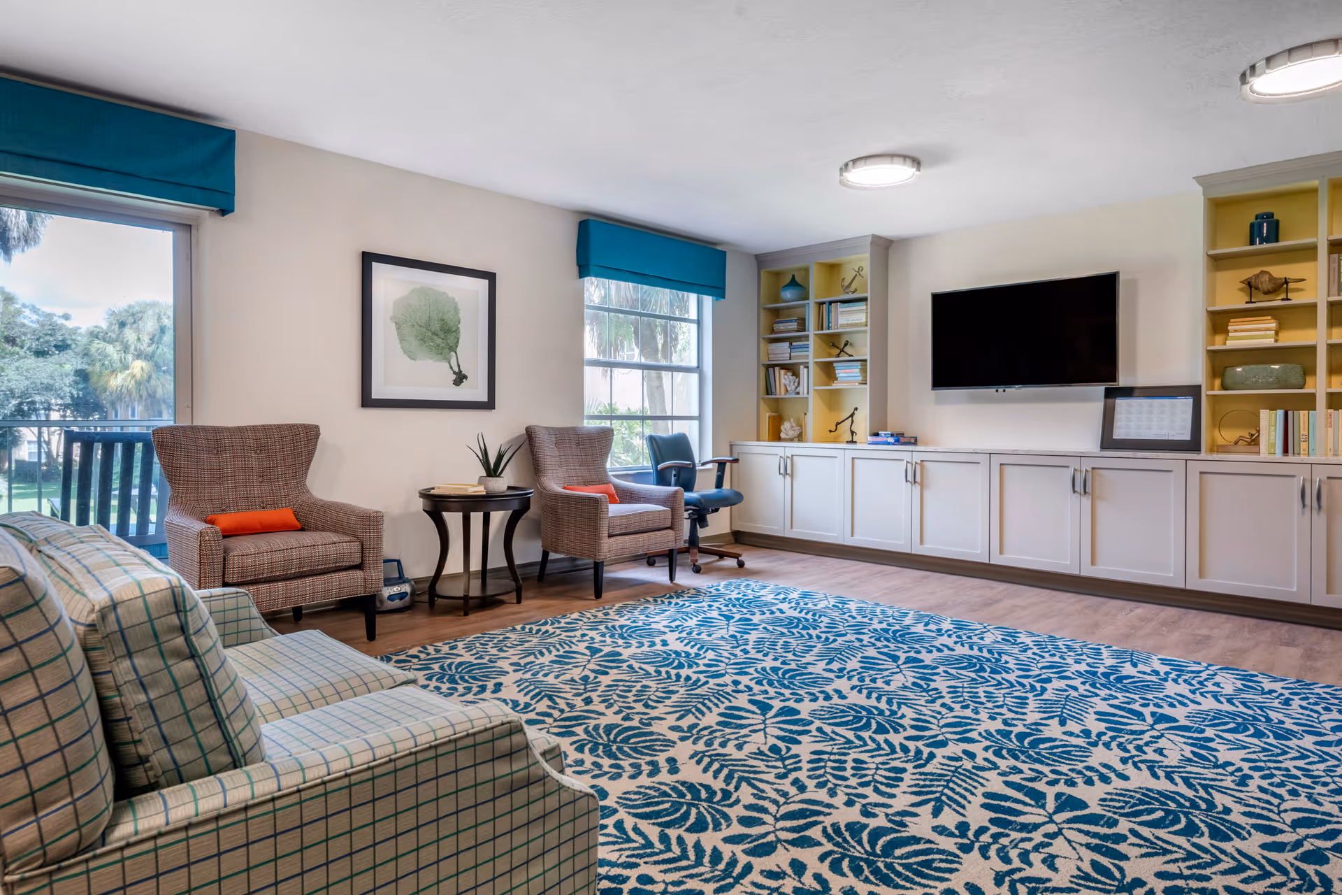 A bright living room with large windows showing outdoor greenery. The room features a patterned blue and white rug, a plaid sofa, two armchairs with orange cushions, a small round side table with a plant, a rolling office chair, built-in white cabinets with yellow shelving, and a wall-mounted flat-screen TV.