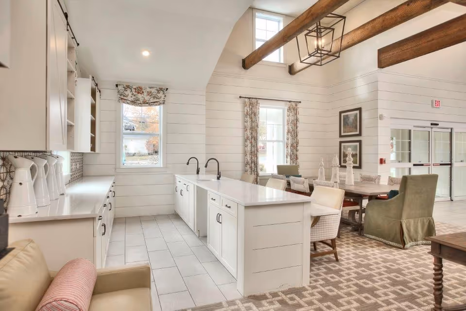 Bright and spacious kitchen area with white cabinetry, two sinks, and a long countertop. The room has large windows with floral curtains, exposed wooden ceiling beams, and a dining area with a wooden table and upholstered chairs. The floor is tiled in the kitchen area and carpeted in the dining area.