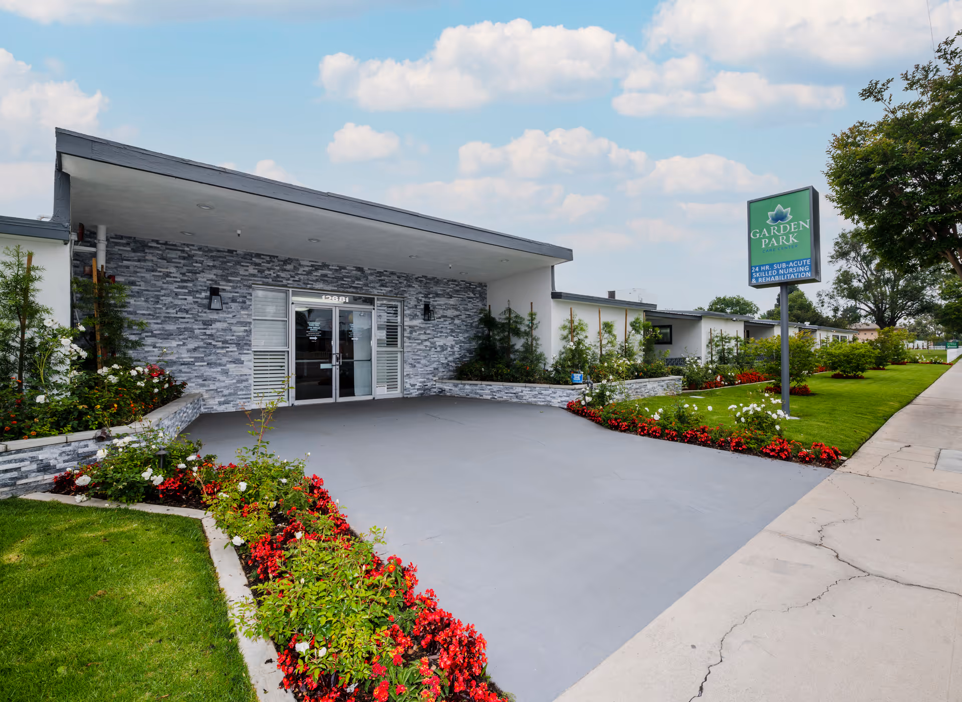 Front exterior view of Garden Park Care Center building with a covered entrance, stone facade, glass double doors, and well-maintained landscaping including green grass and red and white flowers under a partly cloudy sky.