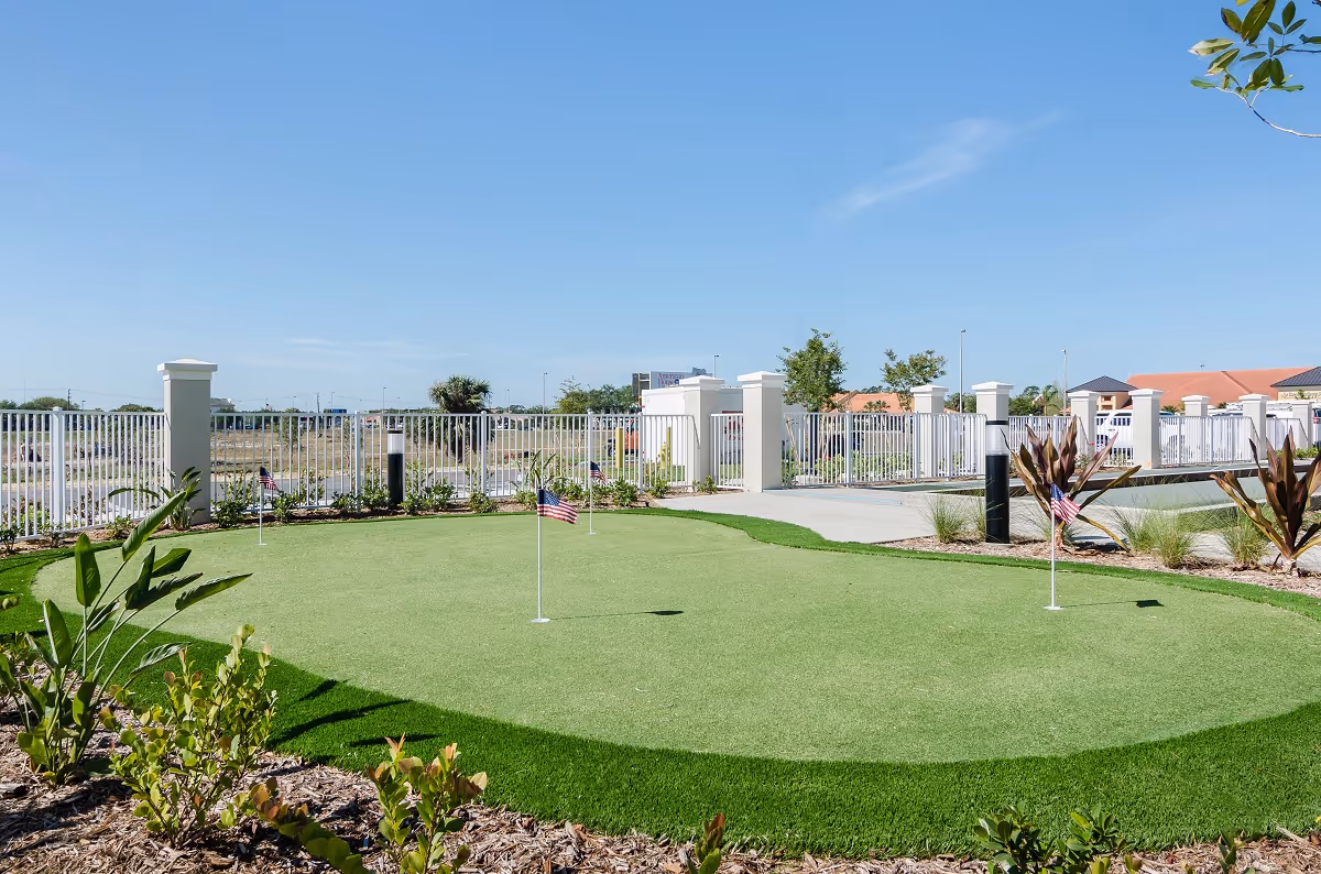 Outdoor putting green with small American flags on the holes, surrounded by plants and a white fence under a clear blue sky at American House Ft. Myers.
