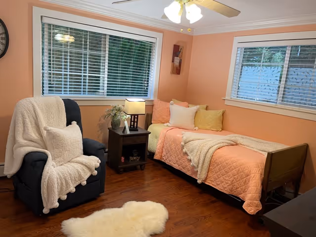 A cozy bedroom with peach-colored walls featuring a single bed with a peach quilt, multiple pillows, and a beige throw blanket. Next to the bed is a dark wooden nightstand with a lamp and a small plant. A comfortable dark armchair draped with a white fluffy blanket and pillow is positioned near the nightstand. The room has two windows with white blinds and wooden floors with a small white fluffy rug.