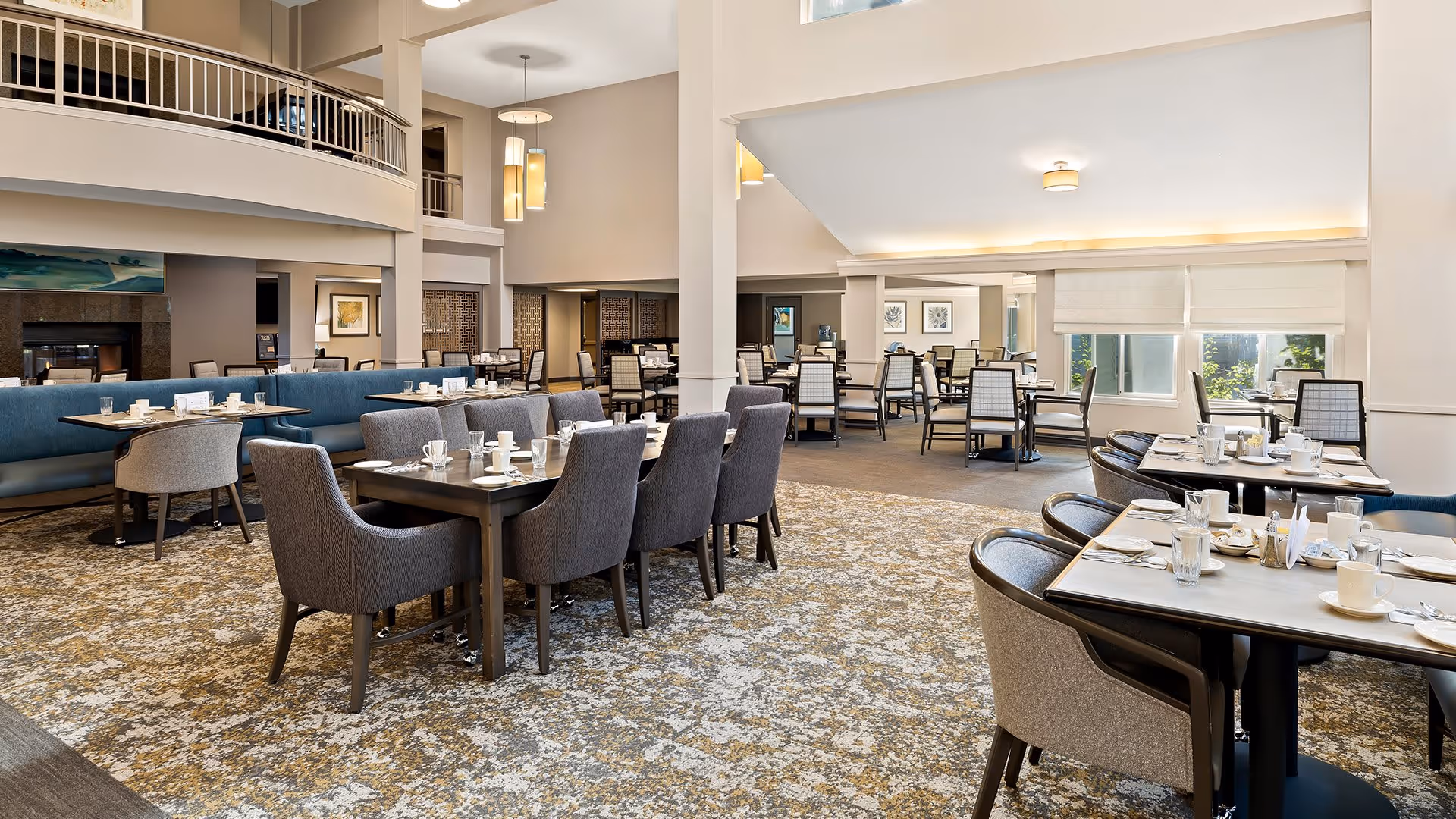 A spacious, well-lit dining room with multiple tables and chairs arranged neatly. The tables are set with cups, glasses, and utensils. The room features a high ceiling, large windows with natural light, and a carpeted floor with a patterned design. There is a second-floor balcony overlooking the dining area.