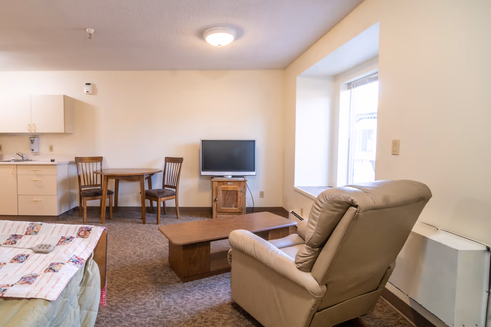 A cozy room with a beige recliner chair facing a wooden coffee table and a small TV on a wooden stand. There is a round wooden table with two chairs near a kitchenette area with cabinets and a sink. A bed with a colorful quilt is partially visible in the foreground. The room has a large window letting in natural light.