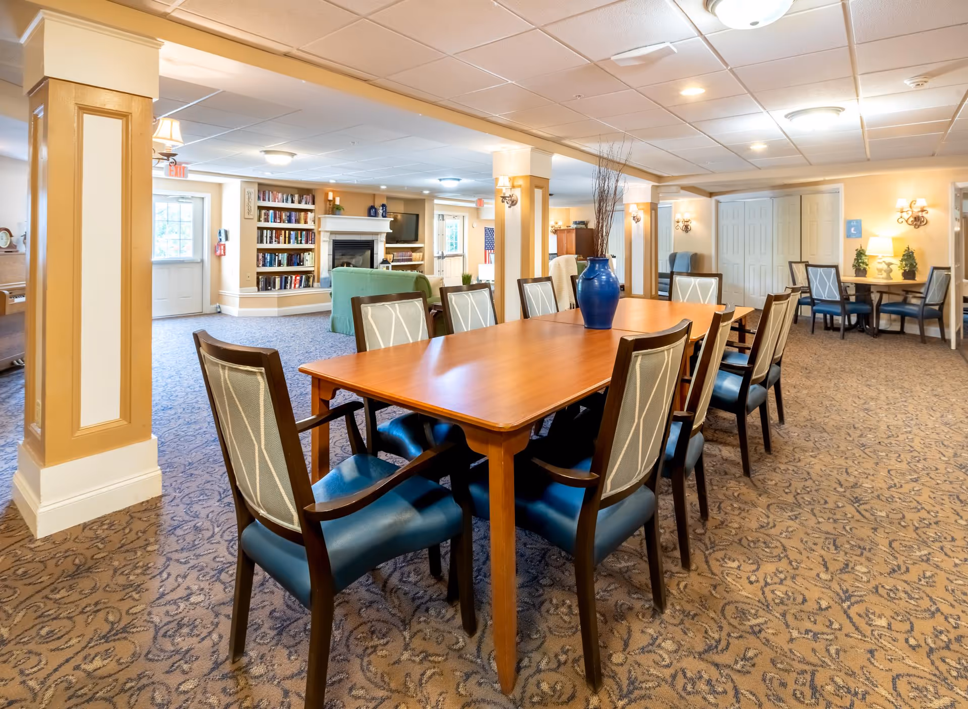 A spacious and well-lit common area in a senior living facility featuring a long wooden table with eight chairs around it. The room has patterned carpet flooring and beige walls with white trim. In the background, there is a cozy seating area with a green sofa, a fireplace, bookshelves filled with books, and a television mounted above the fireplace. Additional seating areas with chairs and small tables are visible further back in the room.