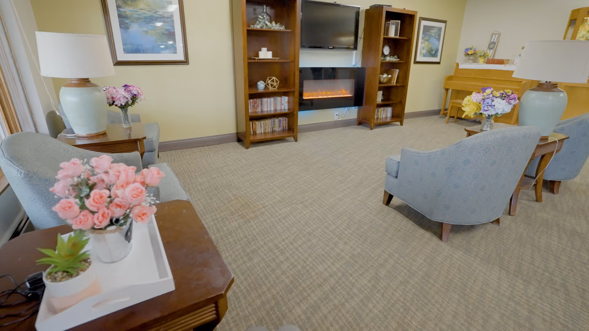 A cozy living room area with light blue upholstered chairs arranged around a wooden coffee table with flower arrangements. There are two large table lamps with light blue bases on side tables. A wall-mounted electric fireplace is flanked by two wooden bookshelves filled with decorative items and DVDs. A piano with a flower vase on top is visible in the background, and framed artwork hangs on the walls.