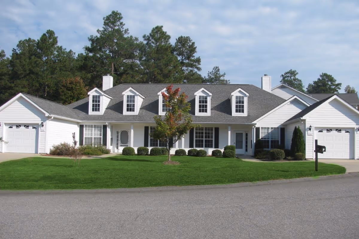 Front exterior view of a white residential building with a gray roof, multiple dormer windows, two garages on either side, a well-maintained green lawn, a small tree in the center, and bushes along the front. The sky is partly cloudy and there are tall pine trees in the background.