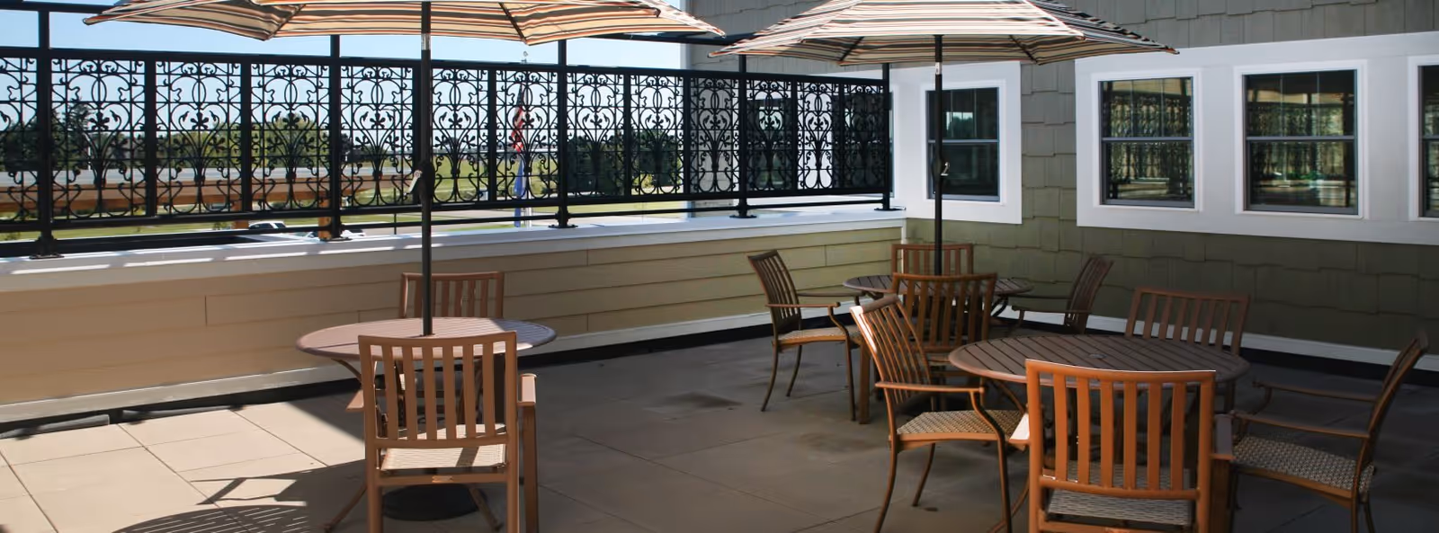 Outdoor patio area with round wooden tables and chairs under large striped umbrellas, surrounded by a decorative black metal railing and adjacent to a building with green siding and white-framed windows.