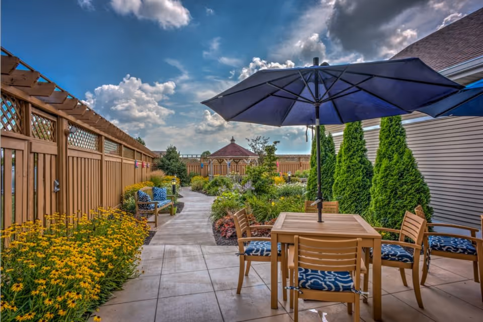 Outdoor patio area with a wooden table and four chairs with blue patterned cushions under a large blue umbrella. A paved walkway leads through a garden with yellow flowers and green shrubs, bordered by a wooden fence on the left and tall evergreen trees on the right. A gazebo is visible in the background under a partly cloudy sky.