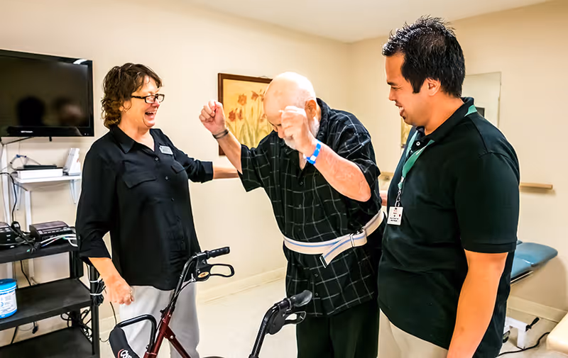 An elderly man using a walker is assisted by two caregivers in a room with medical equipment and a TV mounted on the wall. The man is raising his arms in a celebratory gesture while the caregivers smile and support him.