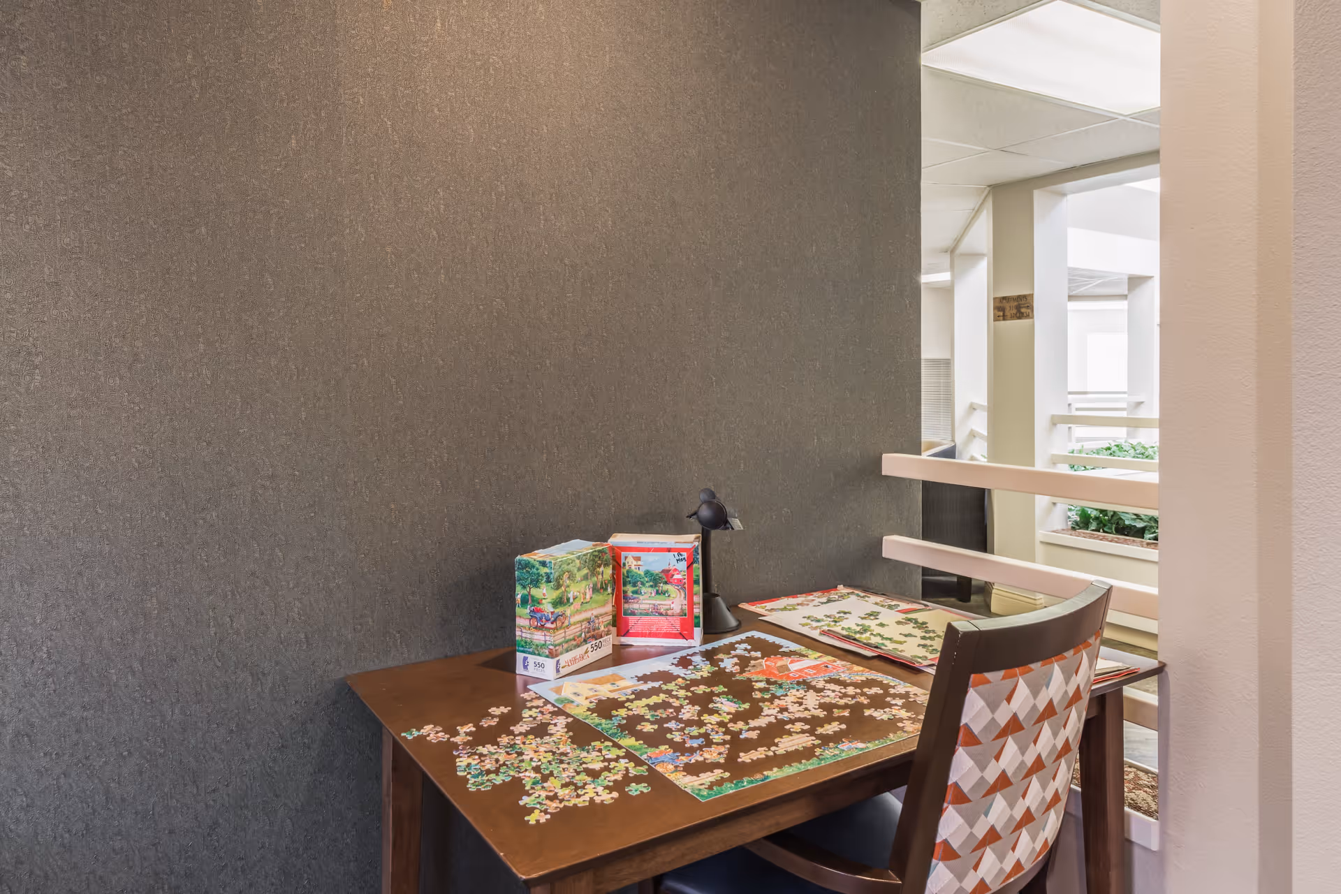 A small wooden table with a partially completed jigsaw puzzle and puzzle pieces scattered on it. There are two puzzle boxes and a small black desk lamp on the table. A chair with a patterned backrest is positioned at the table. The setting appears to be an indoor common area with a gray textured wall and a railing overlooking an open space with natural light and plants.