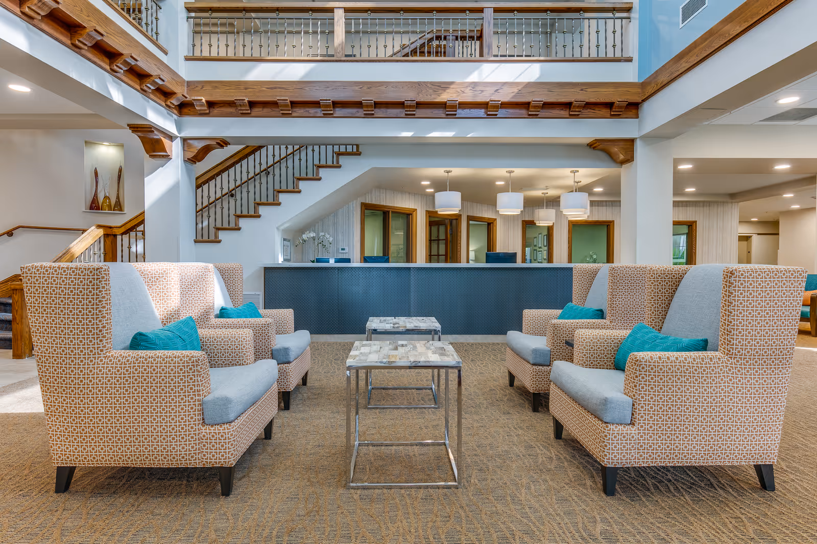 A spacious and well-lit lobby area with four patterned armchairs featuring gray cushions and teal pillows arranged around two small square tables with metal frames and stone tops. Behind the seating area is a reception desk with three pendant lights hanging above it. A staircase with wooden railings is visible on the left side, and the upper floor balcony with wooden trim and metal balusters overlooks the lobby.