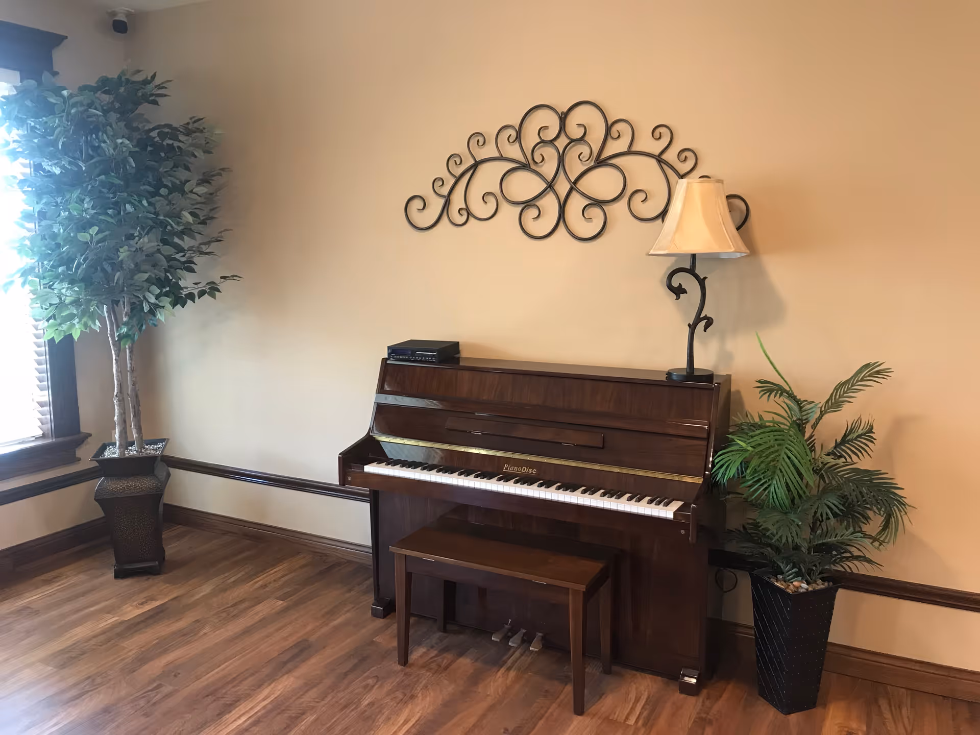 A wooden upright piano with a matching bench is placed against a beige wall. On top of the piano, there is a small electronic device and a decorative lamp with a beige shade. Above the piano, a black ornamental metal wall decoration is mounted. To the left and right of the piano, there are two potted green plants. The floor is wooden, and there is a window with blinds on the left side letting in natural light.