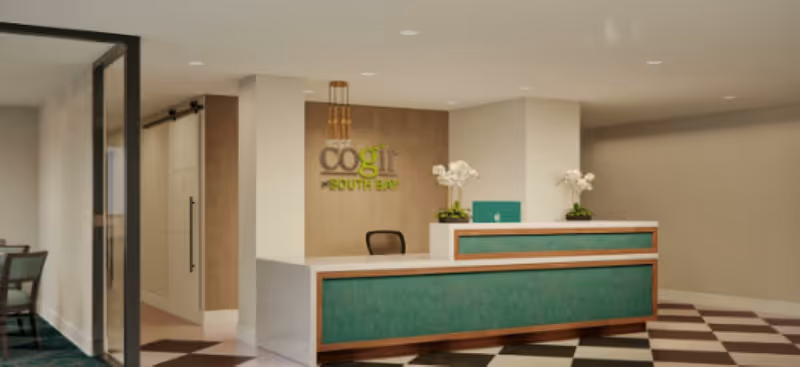 Reception area of Cogir of South Bay featuring a modern front desk with green panels, two white orchid plants, a computer, and a black chair. The background wall displays the facility's name 'cogir SOUTH BAY' in green and gray letters. The floor has a checkered pattern in black and white, and there is a glass door on the left side leading to another room with chairs and a table.