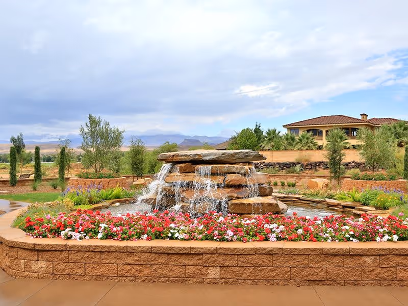 A landscaped outdoor area featuring a tiered stone water fountain surrounded by colorful flowers and greenery, with a building and mountains visible in the background under a partly cloudy sky.