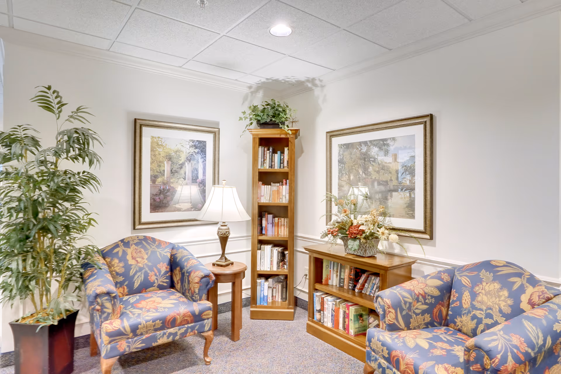 A cozy sitting area with two floral upholstered armchairs, a small wooden side table with a lamp, a tall wooden bookshelf filled with books, and a shorter wooden bookshelf with books and a floral arrangement on top. The walls are decorated with two framed landscape paintings, and there is a potted plant in the corner.