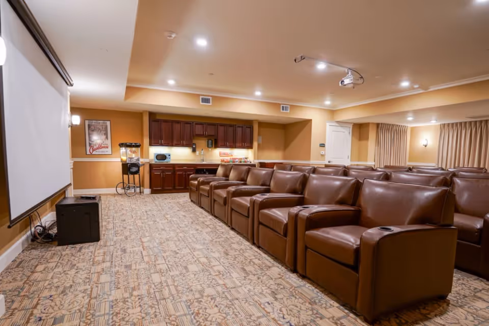 A cozy home theater room with rows of brown leather recliner chairs facing a large projection screen. The room has beige walls, recessed lighting, and a patterned carpet. At the back, there is a kitchenette area with dark wood cabinets, a microwave, a popcorn machine, and a poster on the wall.