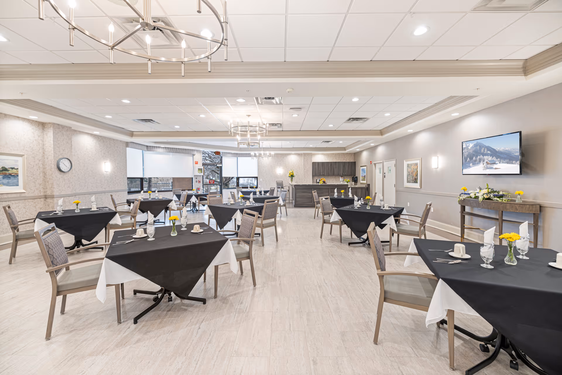 A spacious dining room with several tables covered in black and white tablecloths, each set with glassware, napkins, and small yellow flower centerpieces. The room has light-colored walls, a light wood floor, modern chandeliers, a wall-mounted TV displaying a snowy mountain scene, and large windows letting in natural light.
