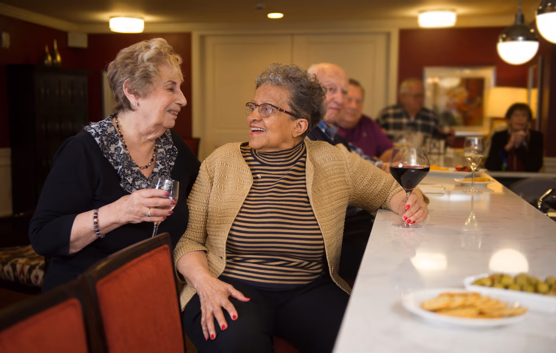Two elderly women sitting at a bar counter in a senior living facility, smiling and holding glasses of wine, with other seniors seated in the background and plates of snacks on the counter.
