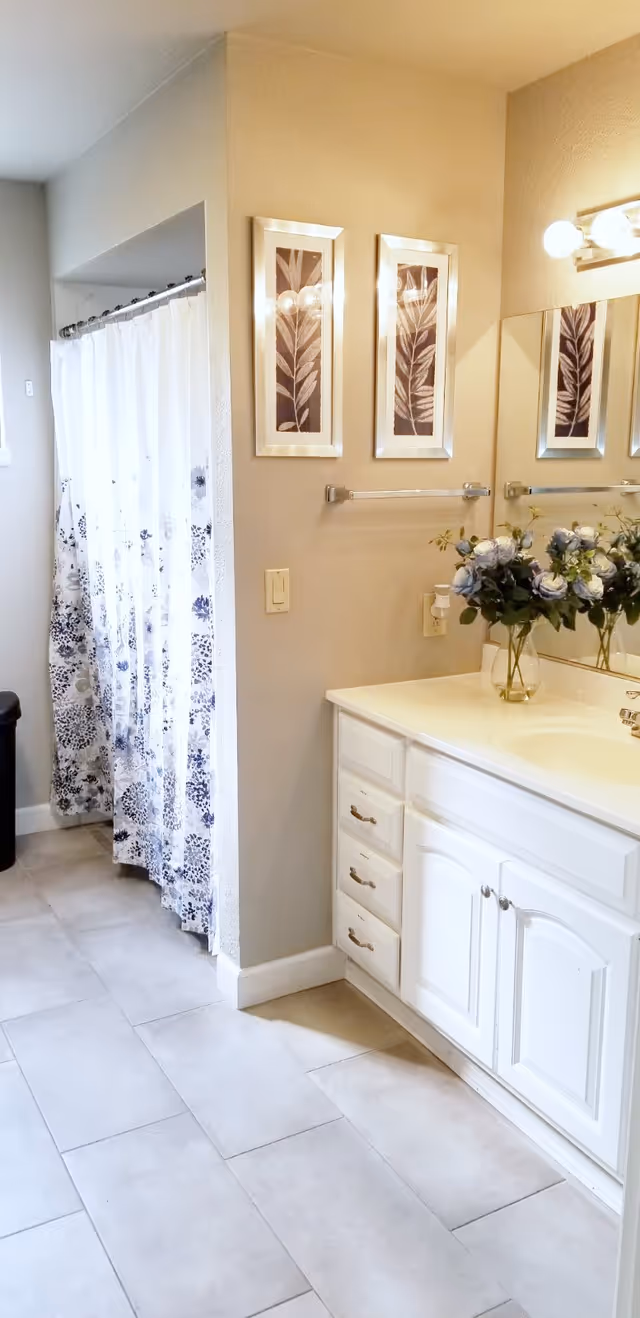 A bathroom with a white vanity cabinet and countertop, a vase with blue and white flowers on the counter, a large mirror above the sink, two framed leaf prints on the wall, a towel rack without a towel, and a shower area with a white and blue floral shower curtain.