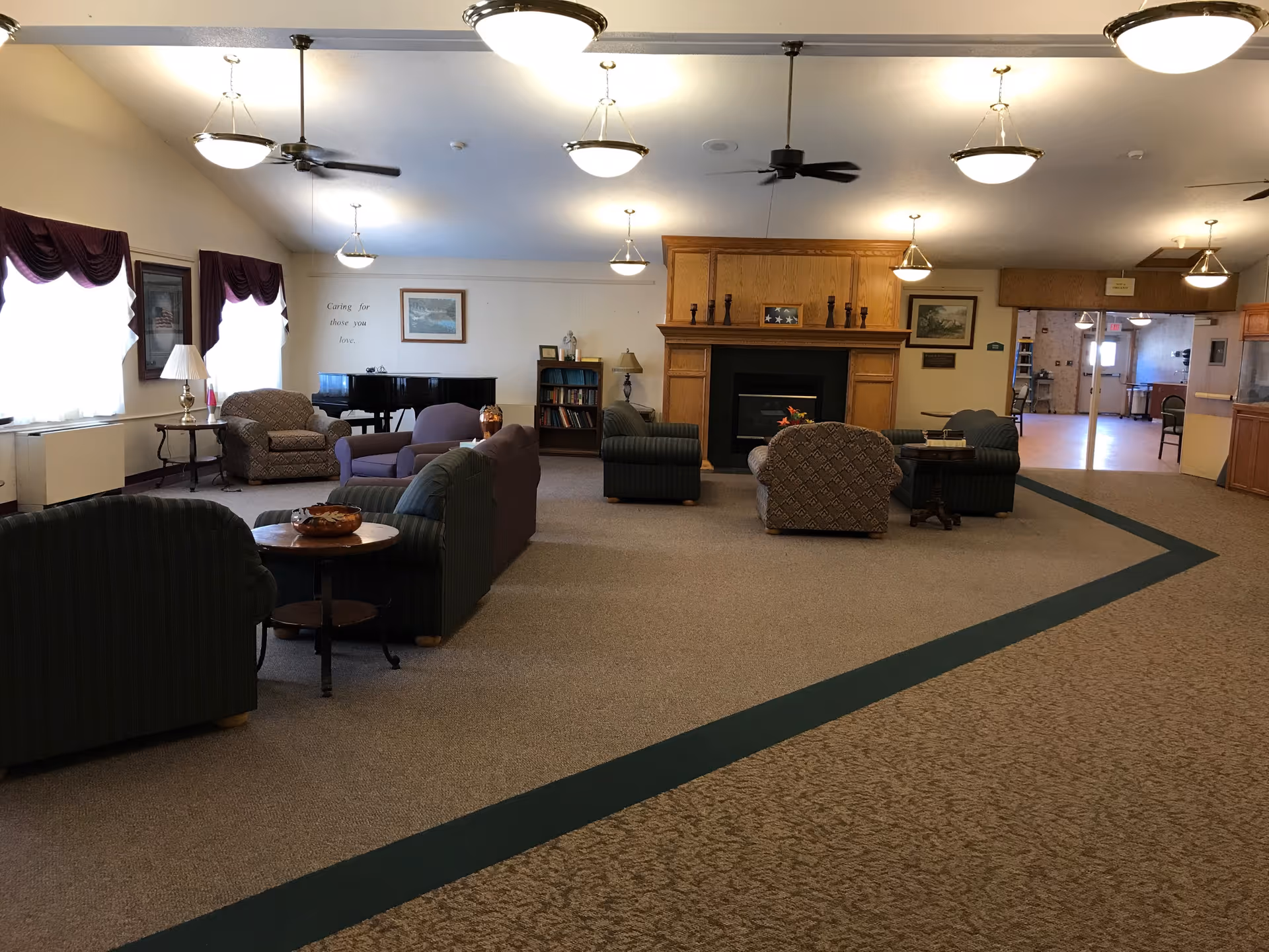 A spacious senior living common area with multiple armchairs and sofas arranged around small tables. There is a fireplace with a wooden mantel in the center of the room, a black grand piano near the windows with purple curtains, and bookshelves against the wall. Ceiling lights and fans are visible, and the room has carpeted flooring with a patterned border.